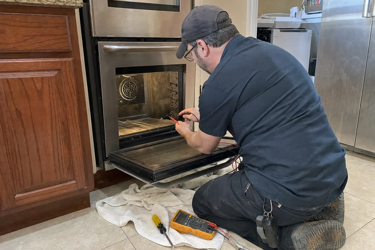 A technician kneeling at a built-in stainless wall oven with the door open, working inside the cavity, a cloth spread under the opening