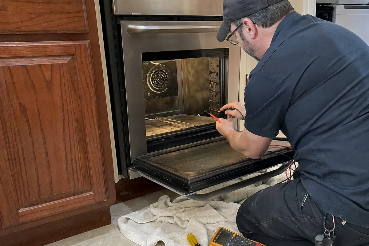 A compact stainless oven set into a run of dark cabinetry, its door open and a hand tool resting on the floor beneath