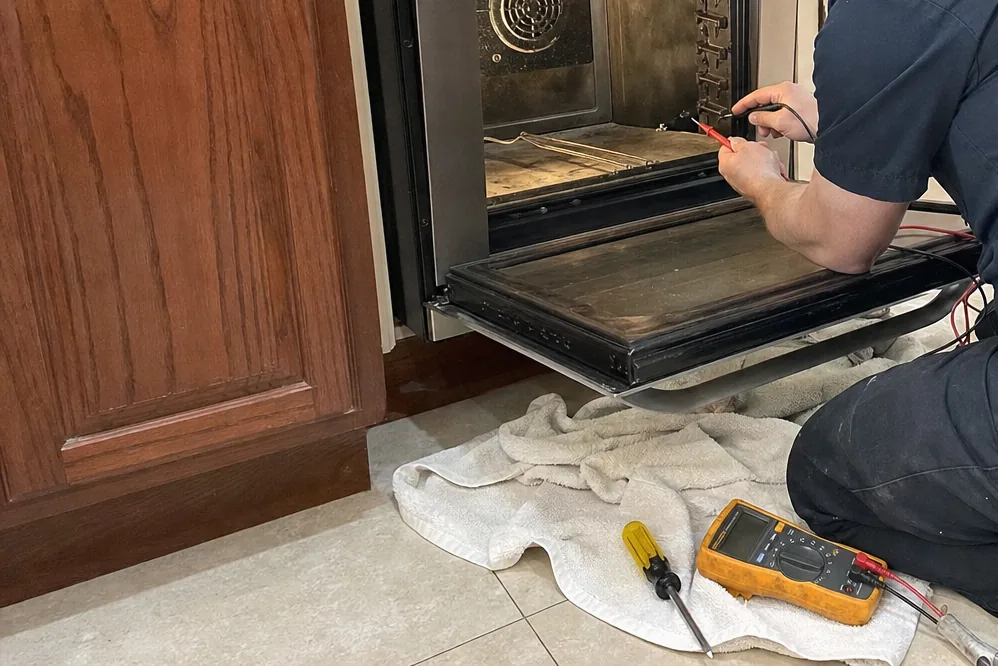 A technician at an open oven, the kitchen behind him bright with light off the window